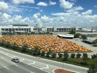 A large parking lot filled with numerous yellow taxis is situated near a large, modern building and an adjacent roadway. The sky is mostly clear with a few scattered clouds, indicating daytime with ample sunlight. The parking lot is surrounded by some greenery and trees, and there are multiple vehicles traveling on the road.