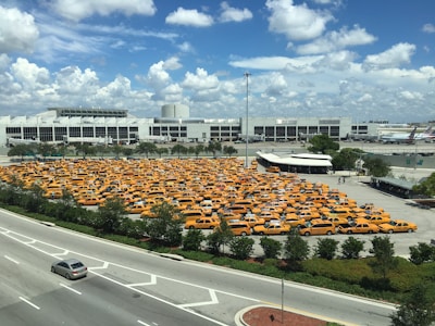 A large parking lot filled with numerous yellow taxis is situated near a large, modern building and an adjacent roadway. The sky is mostly clear with a few scattered clouds, indicating daytime with ample sunlight. The parking lot is surrounded by some greenery and trees, and there are multiple vehicles traveling on the road.