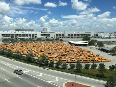 A large parking lot filled with numerous yellow taxis is situated near a large, modern building and an adjacent roadway. The sky is mostly clear with a few scattered clouds, indicating daytime with ample sunlight. The parking lot is surrounded by some greenery and trees, and there are multiple vehicles traveling on the road.