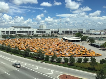 A large parking lot filled with numerous yellow taxis is situated near a large, modern building and an adjacent roadway. The sky is mostly clear with a few scattered clouds, indicating daytime with ample sunlight. The parking lot is surrounded by some greenery and trees, and there are multiple vehicles traveling on the road.