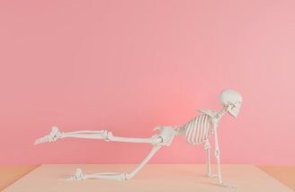 A human skeleton is positioned in a yoga-like pose on a pastel pink and peach background, with one leg extended backward and both arms supporting its body.