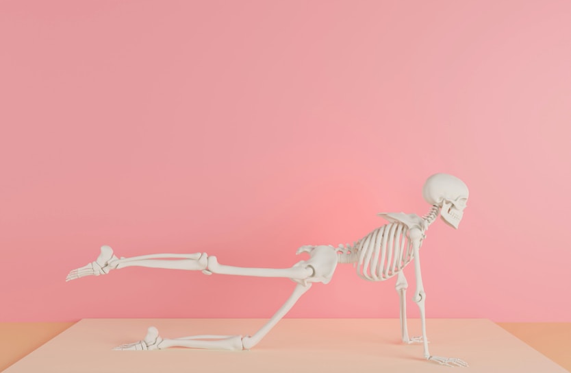A human skeleton is positioned in a yoga-like pose on a pastel pink and peach background, with one leg extended backward and both arms supporting its body.