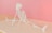 A human skeleton is positioned on a flat surface, performing a wide stretch with legs extended in opposite directions, against a soft pink background. The setting provides a surreal and artistic feel.