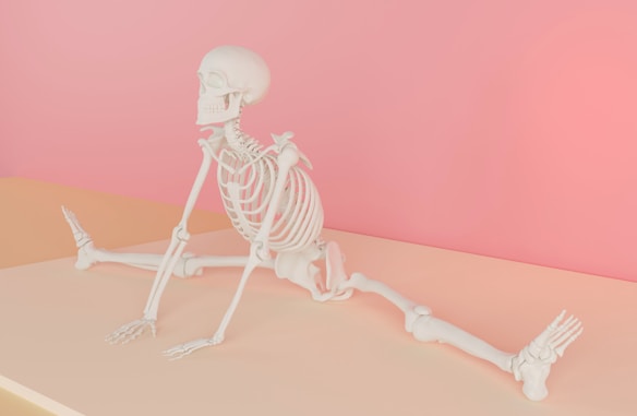 A human skeleton is positioned on a flat surface, performing a wide stretch with legs extended in opposite directions, against a soft pink background. The setting provides a surreal and artistic feel.