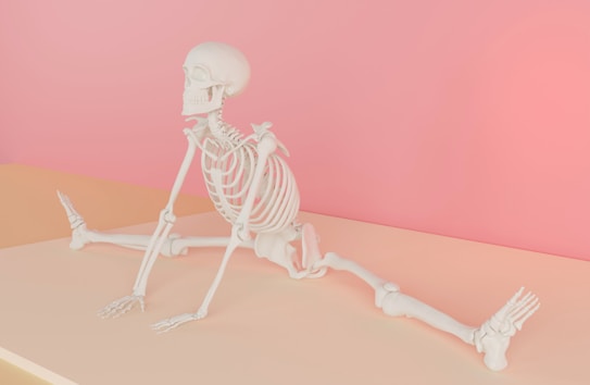 A human skeleton is positioned on a flat surface, performing a wide stretch with legs extended in opposite directions, against a soft pink background. The setting provides a surreal and artistic feel.