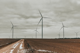 Several tall wind turbines stand in an open field under an overcast sky. The ground is partially covered with patches of snow and a dirt path runs through the scene. The turbines are spaced evenly across the horizon, creating a sense of calm and tranquility.
