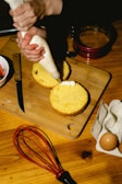 Hands are using a piping bag to add cream filling between two layers of cake on a wooden cutting board. Nearby, there is a serrated knife, an empty cake ring mold, a bowl with something red, an egg carton with eggs, and a whisk.