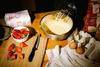 A wooden table is set up for baking with a mixing bowl filled with batter and an electric mixer partially submerged. Fresh strawberries rest on a cutting board beside a knife. A carton holds two eggs, and flour bags are visible in the background.