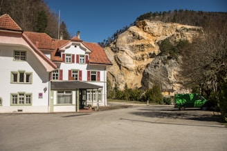 A large, historical-style house with a white facade and red shutters sits in front of a rocky hillside. The roof is covered in reddish-brown tiles. The clear blue sky and surrounding trees add to the serene ambiance. A green truck is parked on the asphalt driveway near the entrance.