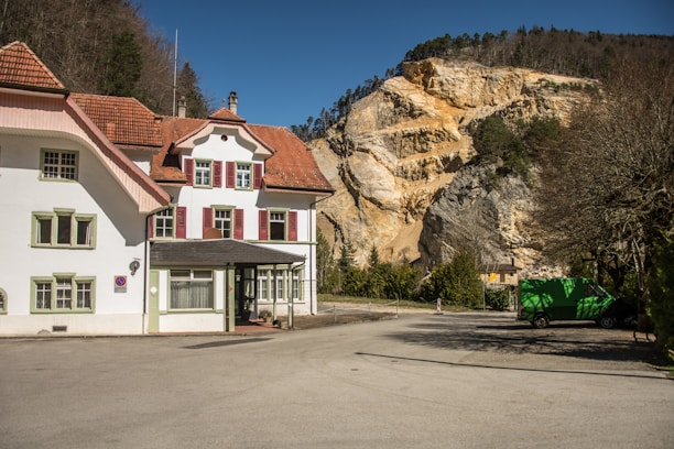 A large, historical-style house with a white facade and red shutters sits in front of a rocky hillside. The roof is covered in reddish-brown tiles. The clear blue sky and surrounding trees add to the serene ambiance. A green truck is parked on the asphalt driveway near the entrance.