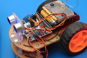 An assembled robot car featuring multiple electronic components, including a circuit board, batteries, sensors, and wires. The car framework is made of light-colored wood, and it has two large black wheels with orange hubs. The background is a solid blue color.
