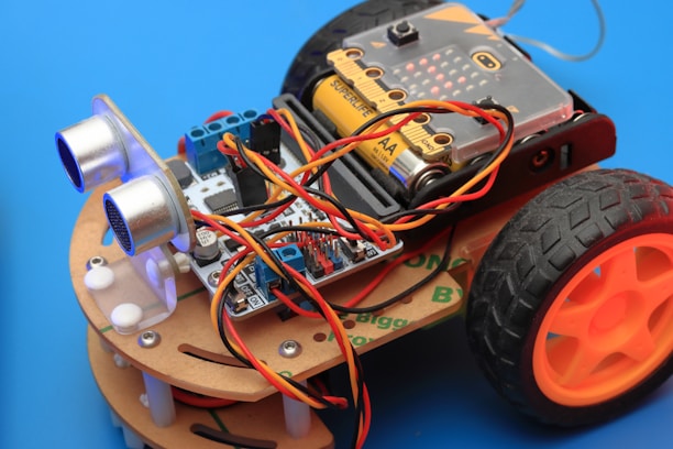 An assembled robot car featuring multiple electronic components, including a circuit board, batteries, sensors, and wires. The car framework is made of light-colored wood, and it has two large black wheels with orange hubs. The background is a solid blue color.