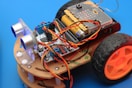An assembled robot car featuring multiple electronic components, including a circuit board, batteries, sensors, and wires. The car framework is made of light-colored wood, and it has two large black wheels with orange hubs. The background is a solid blue color.