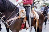 A person wearing traditional cowboy attire is mounted on a dark horse adorned with an ornate saddle. In the background, other riders are visible, also wearing cowboy hats, riding horses in a city setting with trees and buildings.