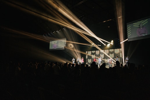 A dimly lit auditorium filled with people in the foreground. On stage, a group of performers or speakers stands in front of a backdrop illuminated by bright, angled stage lights. Two large screens display images on either side of the stage.