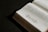 A book lies open on a dark surface, showing pages filled with text on the right side. The left page features a decorative floral design. The pages appear slightly aged, with a sepia tone, and the lighting creates a dramatic shadow effect.
