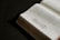 A book lies open on a dark surface, showing pages filled with text on the right side. The left page features a decorative floral design. The pages appear slightly aged, with a sepia tone, and the lighting creates a dramatic shadow effect.