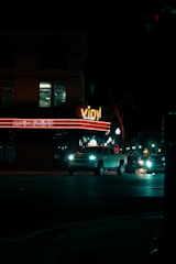 A nighttime city street with a building that has a 'vinyl' sign illuminated in neon lights. The building also displays event names and dates on a lighted marquee. In front of the building, several vehicles, including a white pickup truck, are driving on the road. Street lights line the sidewalk, casting a dim light on the dark street.