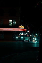 A nighttime city street with a building that has a 'vinyl' sign illuminated in neon lights. The building also displays event names and dates on a lighted marquee. In front of the building, several vehicles, including a white pickup truck, are driving on the road. Street lights line the sidewalk, casting a dim light on the dark street.