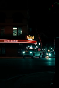 A nighttime city street with a building that has a 'vinyl' sign illuminated in neon lights. The building also displays event names and dates on a lighted marquee. In front of the building, several vehicles, including a white pickup truck, are driving on the road. Street lights line the sidewalk, casting a dim light on the dark street.