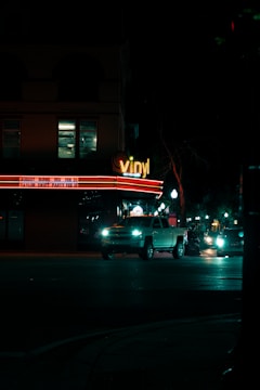 A nighttime city street with a building that has a 'vinyl' sign illuminated in neon lights. The building also displays event names and dates on a lighted marquee. In front of the building, several vehicles, including a white pickup truck, are driving on the road. Street lights line the sidewalk, casting a dim light on the dark street.