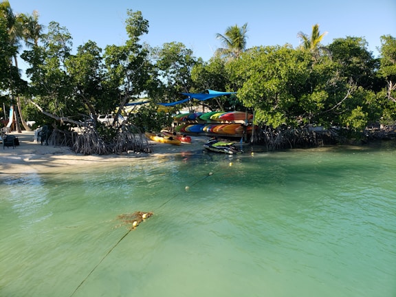 A serene coastal scene features clear turquoise water with a small sandy beach lined by lush green mangroves. Several colorful kayaks are stacked under shade canopies near the shore, and a jet ski is moored nearby.