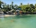 A serene coastal scene features clear turquoise water with a small sandy beach lined by lush green mangroves. Several colorful kayaks are stacked under shade canopies near the shore, and a jet ski is moored nearby.