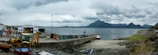 A coastal area featuring a concrete pier extending into a calm body of water. Several boats, including a small red and white boat, are docked by the pier. Various equipment, including a yellow backhoe, litter the area near the shore. The scene is set against a backdrop of distant mountains under a cloudy sky.