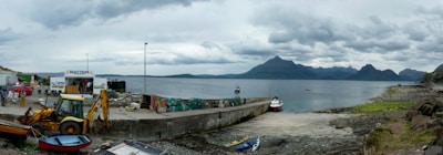A coastal area featuring a concrete pier extending into a calm body of water. Several boats, including a small red and white boat, are docked by the pier. Various equipment, including a yellow backhoe, litter the area near the shore. The scene is set against a backdrop of distant mountains under a cloudy sky.