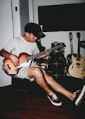 A person plays a bass guitar while seated in a room. The setting includes additional musical instruments like a drum set and two electric guitars. The individual wears casual attire consisting of a t-shirt, shorts, a cap, and sneakers. The room has a relaxed ambiance with wooden flooring.