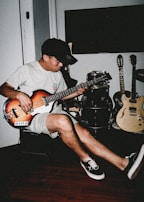 A person plays a bass guitar while seated in a room. The setting includes additional musical instruments like a drum set and two electric guitars. The individual wears casual attire consisting of a t-shirt, shorts, a cap, and sneakers. The room has a relaxed ambiance with wooden flooring.