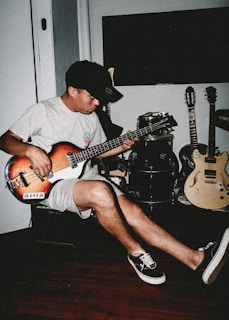 A person plays a bass guitar while seated in a room. The setting includes additional musical instruments like a drum set and two electric guitars. The individual wears casual attire consisting of a t-shirt, shorts, a cap, and sneakers. The room has a relaxed ambiance with wooden flooring.