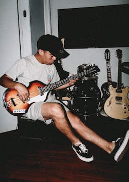 A person plays a bass guitar while seated in a room. The setting includes additional musical instruments like a drum set and two electric guitars. The individual wears casual attire consisting of a t-shirt, shorts, a cap, and sneakers. The room has a relaxed ambiance with wooden flooring.