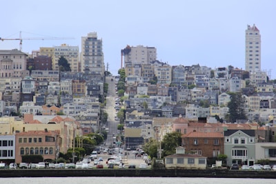 A dense urban area of San Francisco featuring a mix of historical and modern architecture. There are multi-story buildings, townhouses, and green spaces interspersed between the structures. The photo captures a hilly street lined with parked cars and trees, leading up to a cluster of buildings. On the left, a large crane is visible, indicating ongoing construction.