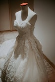 A mannequin is adorned with an elegant white wedding dress featuring intricate lace and embroidery. The dress has a strapless bodice and a full skirt, creating a classic and sophisticated bridal look.