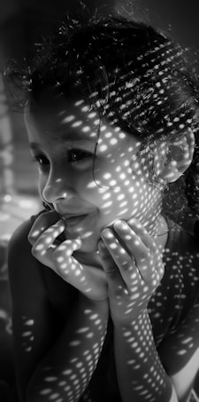 A monochrome portrait of a young girl with a thoughtful expression, her hands gently resting on her chin. Dappled shadows create intricate patterns on her face, likely caused by light filtering through a perforated surface.