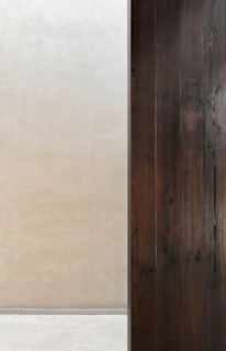 A minimalist composition features a stark contrast between two surfaces. On the left, there is a smooth, light beige wall, while on the right, a series of dark wooden panels provide texture and depth. The two areas are divided by a thin vertical line, creating a clear separation. Below, a simple concrete floor completes the setting.