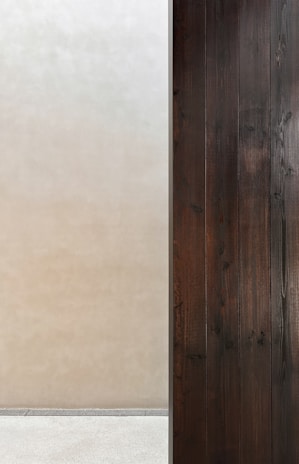 A minimalist composition features a stark contrast between two surfaces. On the left, there is a smooth, light beige wall, while on the right, a series of dark wooden panels provide texture and depth. The two areas are divided by a thin vertical line, creating a clear separation. Below, a simple concrete floor completes the setting.