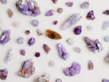 Various crystals and gemstones are scattered across a light background. The stones are predominantly purple, with shades ranging from amethyst to lighter lavender hues. Some stones exhibit sparkling facets, with a mix of raw and polished textures. A few stones also display brown and clear colors.
