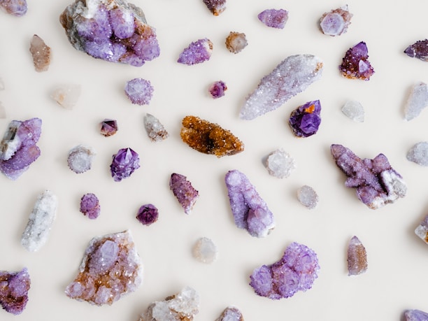 Various crystals and gemstones are scattered across a light background. The stones are predominantly purple, with shades ranging from amethyst to lighter lavender hues. Some stones exhibit sparkling facets, with a mix of raw and polished textures. A few stones also display brown and clear colors.