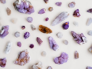 Various crystals and gemstones are scattered across a light background. The stones are predominantly purple, with shades ranging from amethyst to lighter lavender hues. Some stones exhibit sparkling facets, with a mix of raw and polished textures. A few stones also display brown and clear colors.
