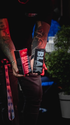 A person with tattooed arms is holding two rectangular product boxes, one primarily red labeled 'Ultra' and the other primarily black labeled 'Black'. The person is wearing a black shirt, and a lanyard with a blue pattern is visible. In the background, there is a blurred blue chair and a green potted plant.