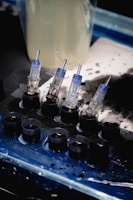 A set of small ink caps filled with black ink are arranged in a row on a blue and black protective sheet. Nearby are several transparent cartridges with needles, likely used for a tattoo machine. The background includes blurred containers and splatters of black ink.