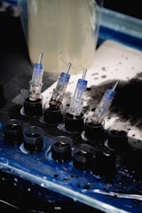A set of small ink caps filled with black ink are arranged in a row on a blue and black protective sheet. Nearby are several transparent cartridges with needles, likely used for a tattoo machine. The background includes blurred containers and splatters of black ink.