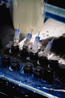 A set of small ink caps filled with black ink are arranged in a row on a blue and black protective sheet. Nearby are several transparent cartridges with needles, likely used for a tattoo machine. The background includes blurred containers and splatters of black ink.