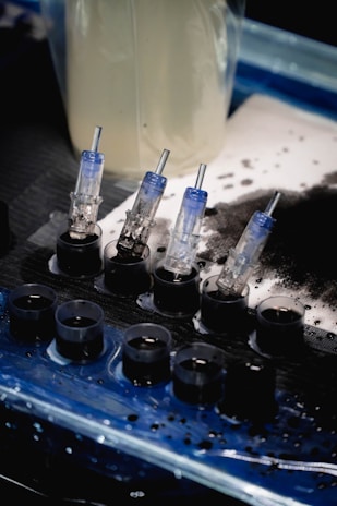 A set of small ink caps filled with black ink are arranged in a row on a blue and black protective sheet. Nearby are several transparent cartridges with needles, likely used for a tattoo machine. The background includes blurred containers and splatters of black ink.