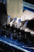 A set of small ink caps filled with black ink are arranged in a row on a blue and black protective sheet. Nearby are several transparent cartridges with needles, likely used for a tattoo machine. The background includes blurred containers and splatters of black ink.