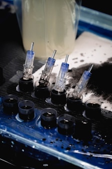 A set of small ink caps filled with black ink are arranged in a row on a blue and black protective sheet. Nearby are several transparent cartridges with needles, likely used for a tattoo machine. The background includes blurred containers and splatters of black ink.