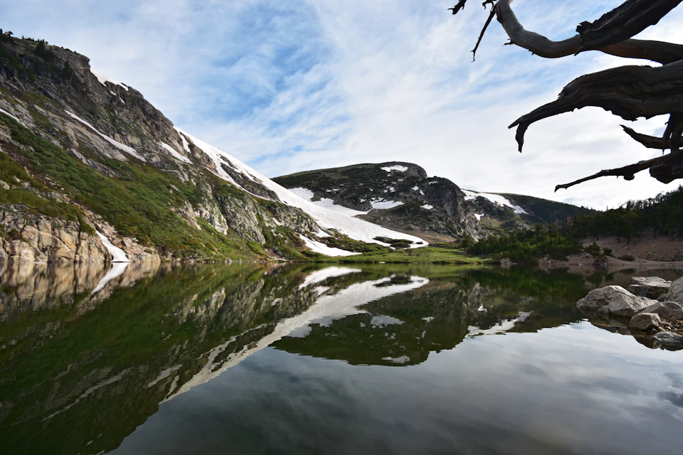 The image depicts a serene mountain lake nestled amidst lush green mountains and trees. The calm, still waters of the lake reflect the surrounding landscape, creating a mirror-like effect that enhances the beauty of the scene. On one side of the lake, there is a large rock formation with patches of snow on its surface, adding to the picturesque setting. A tree branch extends from this rocky outcropping towards the right side of the image, providing a natural frame for the view. The sky above is a clear blue, dotted with fluffy white clouds that add depth and dimension to the scene.