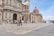 A historic church with intricate stone carvings stands prominently. In front of the church, people are walking on a spacious stone plaza. The sky is clear and blue.